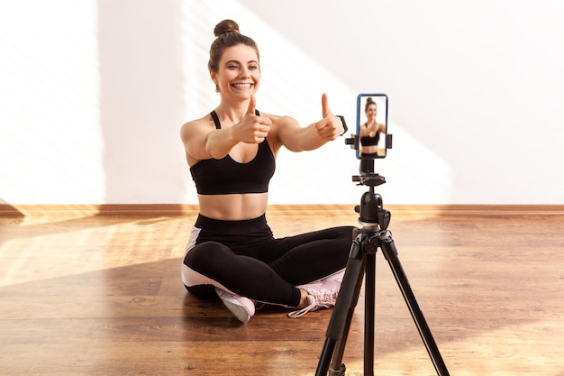 L'allenatore di fitness positivo mostra i pollici in su alla fotocamera del telefono, gioendo del buon allenamento online con i follower, indossando top e collant sportivi neri. Colpo in studio a figura intera illuminato dalla luce solare dalla finestra.