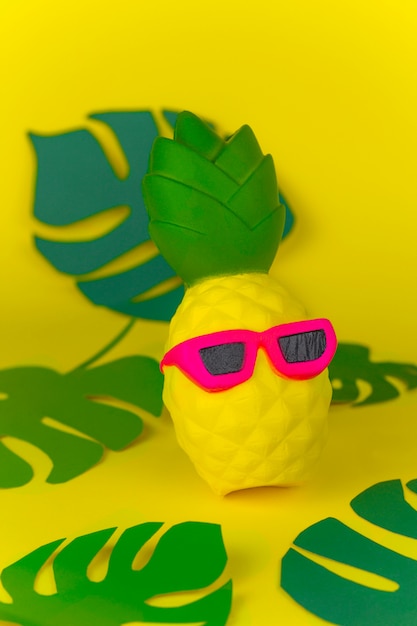 L&#39;ananas squishy del giocattolo in occhiali da sole su fondo giallo fra la carta ha tagliato le foglie tropicali