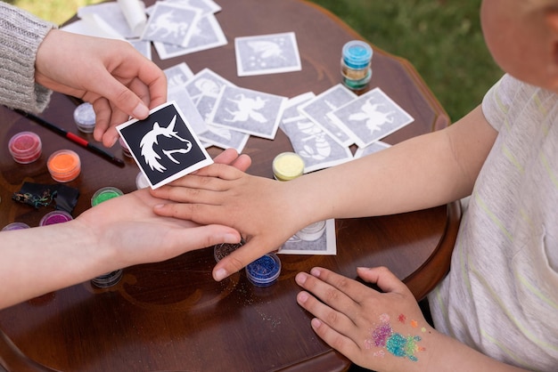 L'animatore per bambini sta creando un tatuaggio luccicante scintillante sulla mano di un bambino a una festa di compleanno