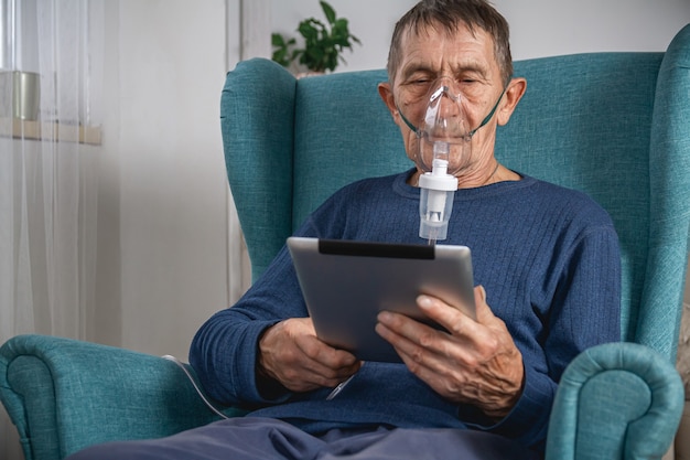 L'anziano anziano si siede su una poltrona con una maschera ad ossigeno e un tablet