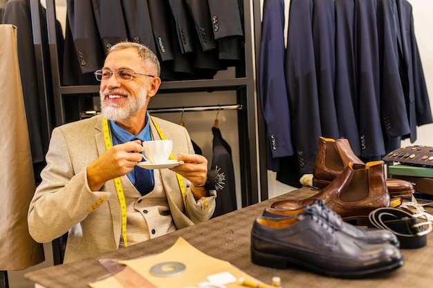 L'anziano bell'uomo con la barba grigia indossa un bel vestito che si prende una pausa dal cucire con una tazza di caffè allo studio dell'atelier