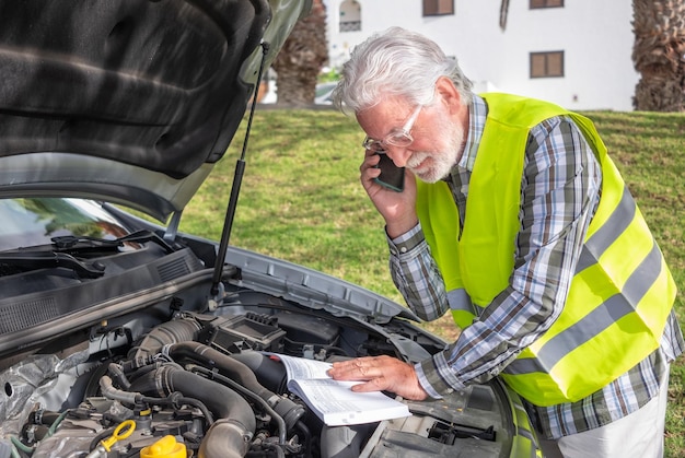 L'anziano conducente si è fermato in strada per un guasto all'auto apre il cofano e parla al telefono con il meccanico Legge il libretto di istruzioni per capire cosa si è rotto