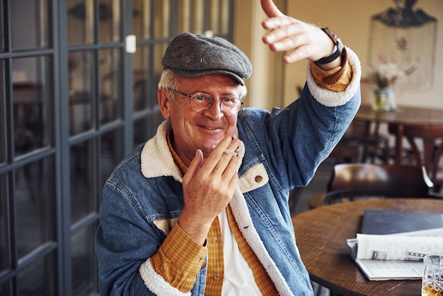 L'anziano elegante in abiti alla moda e con gli occhiali si siede al bar con sigaretta e parla.