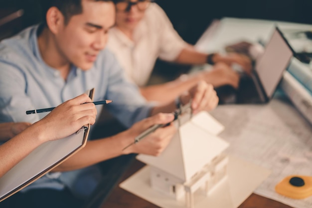 L'architetto maschio passa la misurazione e la realizzazione di una casa modello con loro squadra sulla scrivania al tramonto. Ingegnere, ingegneria, architettura, design, pianificazione, concetto di occupazione.