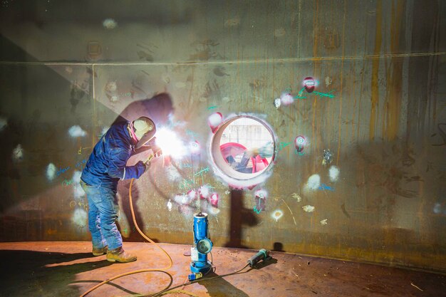 L'arco metallico del lavoratore maschio della saldatura fa parte del serbatoio di stoccaggio del petrolio e del gas del serbatoio del macchinario della costruzione del tombino del macchinario all'interno confinato