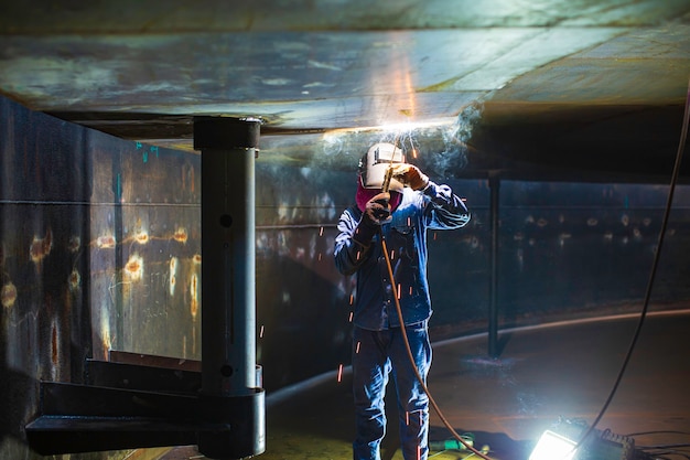 L'arco metallico del lavoratore maschio di saldatura fa parte del serbatoio di stoccaggio di petrolio e gas per la costruzione di condutture di serbatoi di macchinari all'interno di spazi ristretti.