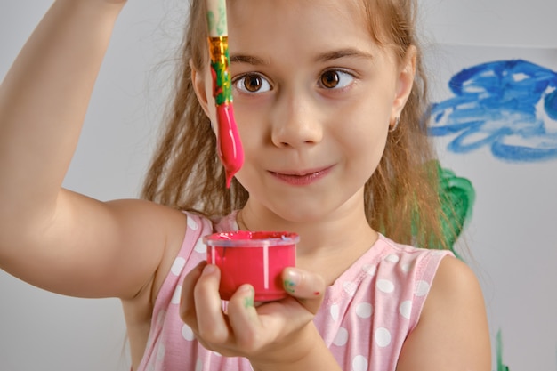 L'artista della bambina in un vestito rosa è in piedi dietro il cavalletto e dipinge con il pennello su tela