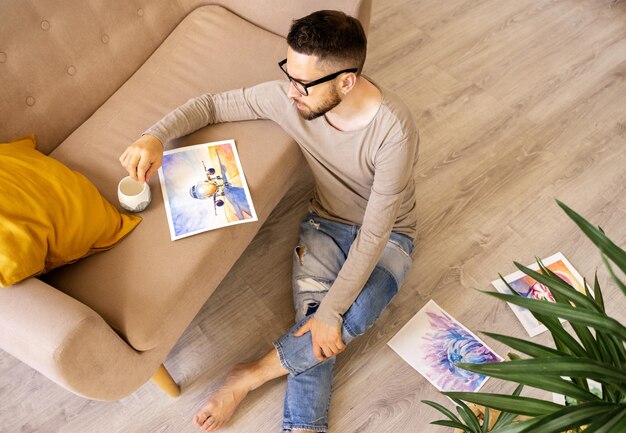 L'artista è seduto per terra con una tazza di caffè guarda un disegno con un aeroplano è triste e sogna di volare in vacanza durante una pandemia Emozioni vere lifestyle