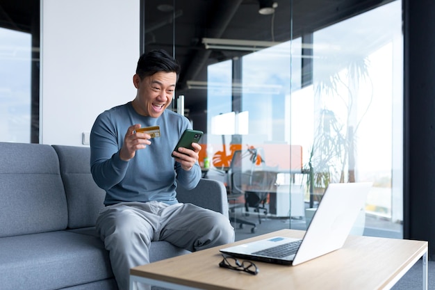 L'asiatico di successo e felice effettua acquisti online con una carta di credito e un'applicazione sul telefono un uomo d'affari trascorre del tempo in un ufficio moderno su un laptop