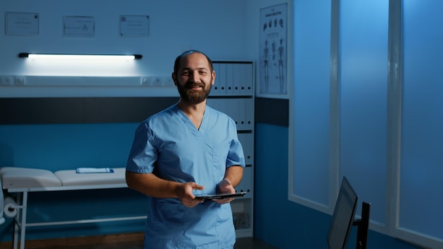 L'assistente dell'uomo caucasico che tiene il computer tablet digitando la perizia medica controllando il rapporto sulla malattia del paziente. Infermiera in uniforme blu che lavora fuori orario nell'ufficio dell'ospedale. Servizio e concetto di assistenza sanitaria