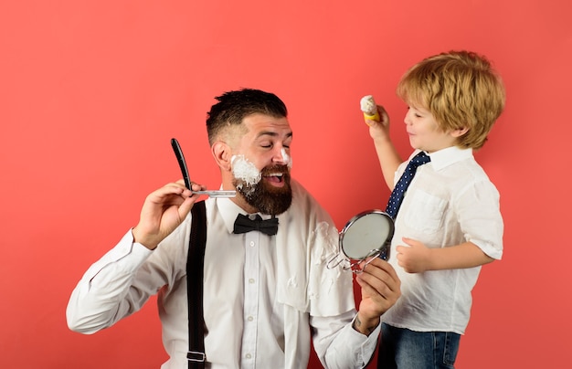 L'assistente della festa del papà per il bambino di papà tiene lo specchio per il barbiere del padre che rade l'uomo barbuto in