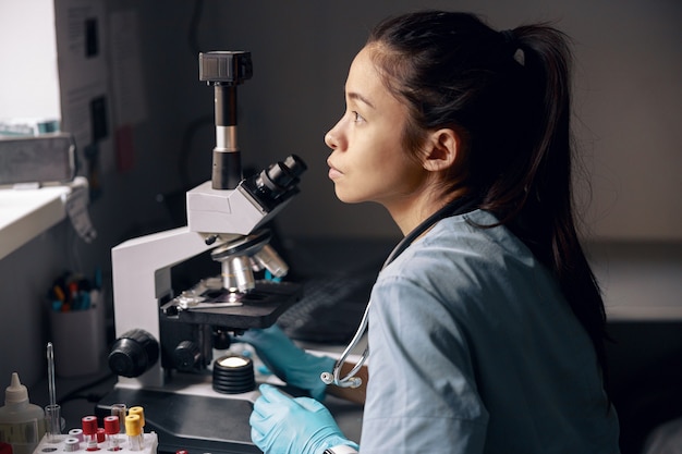 L'assistente di laboratorio asiatico premuroso si siede vicino al microscopio guardando fuori dalla finestra sul posto di lavoro