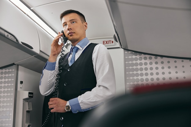 L'assistente di volo parla a bordo del telefono nella cabina passeggeri del jet aereo. Interni aereo moderno. Vista dal basso di un uomo multirazziale indossa l'uniforme. Aviazione civile commerciale. Concetto di viaggio aereo