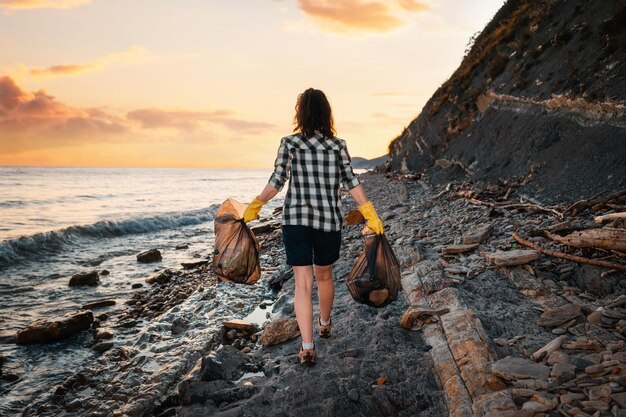 L'attivista donna cammina lungo la costa selvaggia e porta due sacchi di spazzatura sullo sfondo l'oceano e il tramonto Il concetto di pulizia della spiaggia dall'inquinamento