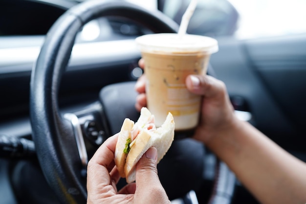 L'autista di una donna asiatica tiene una tazza di caffè ghiacciata e un panino per mangiare e bere in auto pericoloso e rischiare un incidente