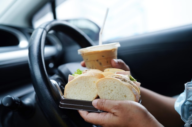 L'autista di una donna asiatica tiene una tazza di caffè ghiacciata e un panino per mangiare e bere in auto pericoloso e rischiare un incidente