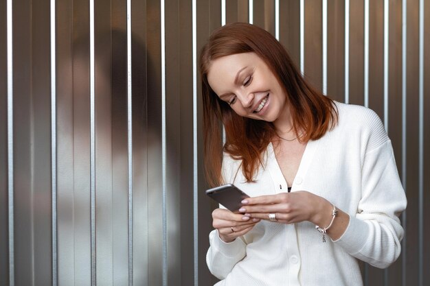 L'economista femminile europeo di successo positivo effettua il pagamento di denaro sul sito Web pone con il telefono cellulare vestito con un abito bianco sorride felicemente invia un messaggio di testo gode della comunicazione online