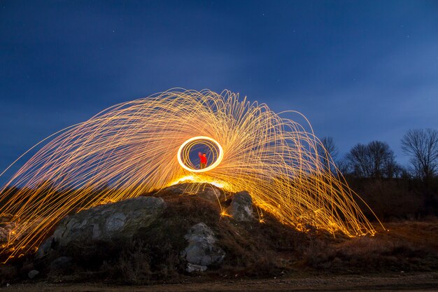 L'esposizione lunga ha sparato dell'uomo che sta sulla collina rocciosa che fila la lana d'acciaio nel cerchio che fa le docce del fuoco d'artificio delle scintille d'ardore gialle luminose sul fondo blu del cielo notturno. Concetto di arte della pittura leggera.