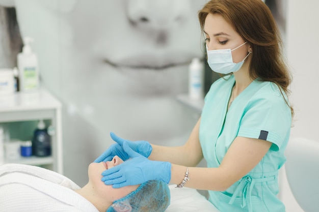 L'estetista della giovane donna in maschera applica il gel di contatto sulla gamba di una donna per la depilazione laser dentro