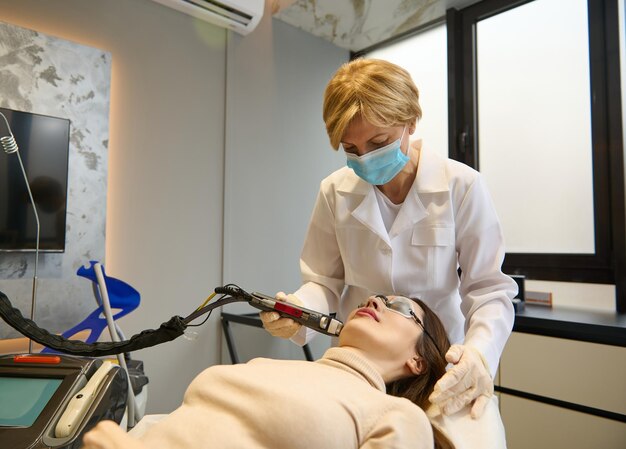 L'estetista fiduciosa esegue procedure cosmetiche sul viso di una donna utilizzando un laser modale per rimuovere le macchie dell'età e dell'età