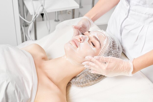 L'estetista massaggia la maschera crema sulla pelle del viso femminile per la procedura di ringiovanimento nel salone di bellezza