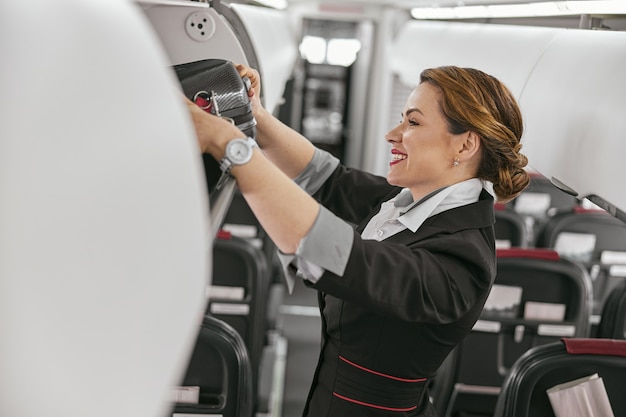 L'hostess prende i bagagli dallo scaffale nella cabina passeggeri del jet aereo. Interni aereo moderno. Uniforme europea sorridente di usura della donna. Aviazione civile commerciale. Concetto di viaggio aereo