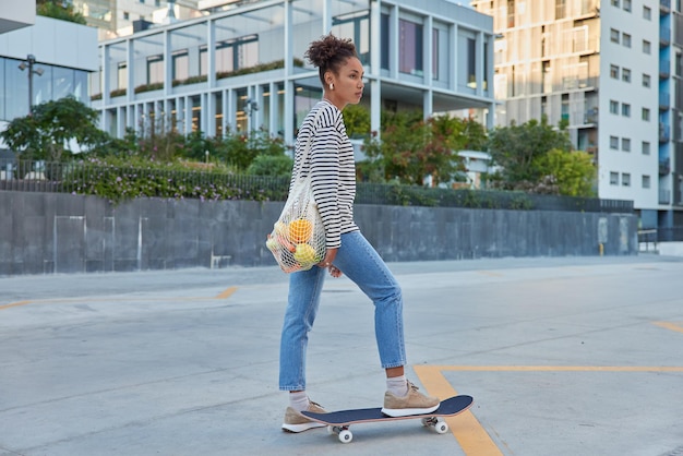 L'immagine a figura intera di una bella ragazza attiva cavalca lo skateboard in città ritorna dal mercato indossa jeans a righe e scarpe da ginnastica porta una borsa a rete con verdure pone all'aperto in ambiente urbano