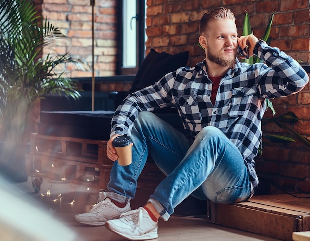 L'immagine completa del corpo di un uomo biondo alla moda con la barba beve il caffè e utilizza lo smartphone in una stanza con interni soppalcati.