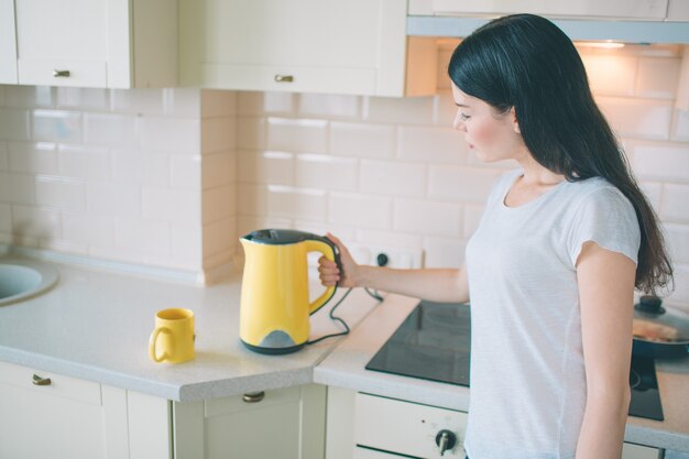 L'immagine dei supporti del brunette e osserva sul bollitore. Lei lo accende. La donna tiene la mano sul lato del bollitore. Lei è in cucina.