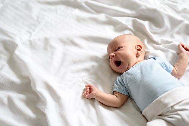 L'immagine del bambino in tuta blu e pantaloni bianchi si trova a sbadigliare sul letto bianco con lo spazio della copia