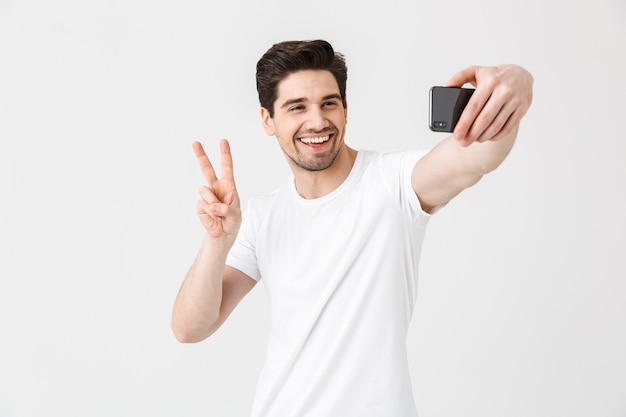 L'immagine del giovane emotivo felice che posa isolato sopra la parete bianca che mostra il gesto di pace prende il selfie dal telefono cellulare.