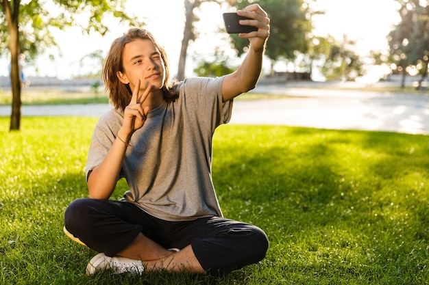 L'immagine del giovane ragazzo allegro che si siede sull'erba nel parco usando il telefono prende un selfie.