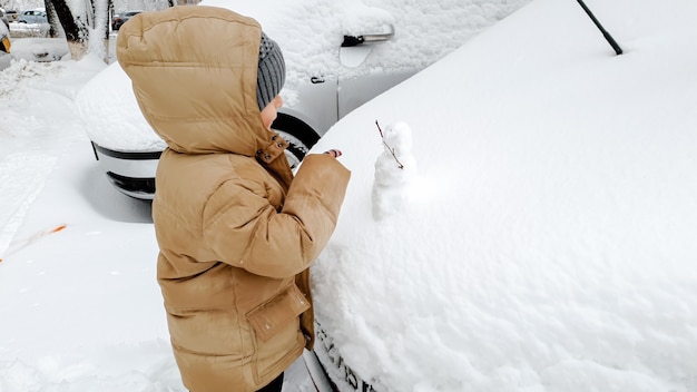 L'immagine del ragazzo ripulisce l'auto coperta di neve dopo la caduta della neve. Nella fredda mattina d'inverno