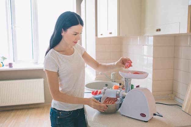 L'immagine della donna sta e tiene il pezzo di pomodoro sopra il tritacarne. Tiene la ciotola con carne di forza. La donna è seria e concentrata.