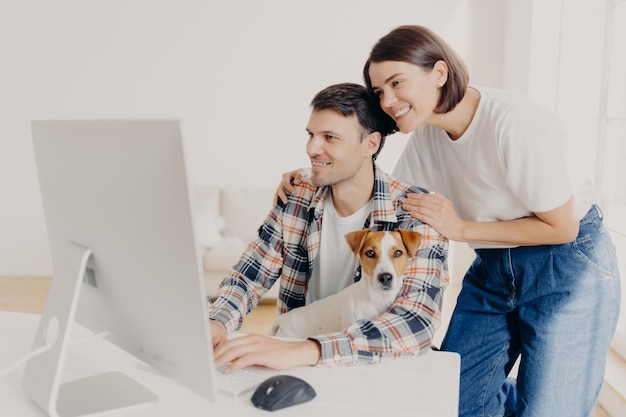 L'immagine della donna tocca delicatamente le spalle dei mariti guardano positivamente nel monitor del computer naviga in Internet acquista mobili online per il loro nuovo appartamento L'uomo felice lavora su un dispositivo moderno con il cane