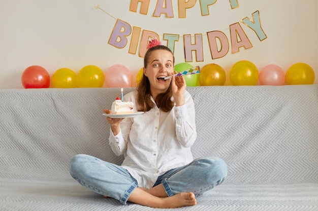 L'immagine della giovane donna attraente soddisfatta felice che indossa una camicia bianca che celebra il compleanno seduta da sola sul divano con il corno del partito che soffia la torta sembra estremamente felice