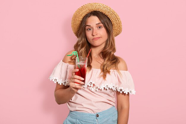 L'immagine della giovane donna che indossa la camicia estiva e cappello di paglia, tenendo in mano un bicchiere di dolce cocktailin mano, sembra annoiato, esprime tristezza, posa di modello isolato sulla parete di studio rosa. Concetto di emozioni di persone.