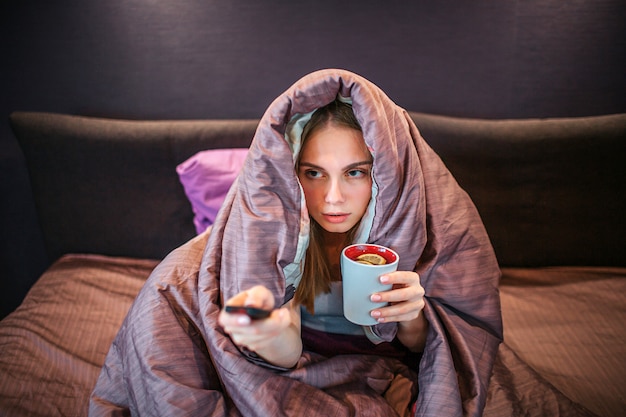 L'immagine della giovane donna coperta di coperta spessa e guarda la TV. Tiene il telecomando in una mano e una tazza di tè con un pezzo di limone in un'altra. La ragazza è malata