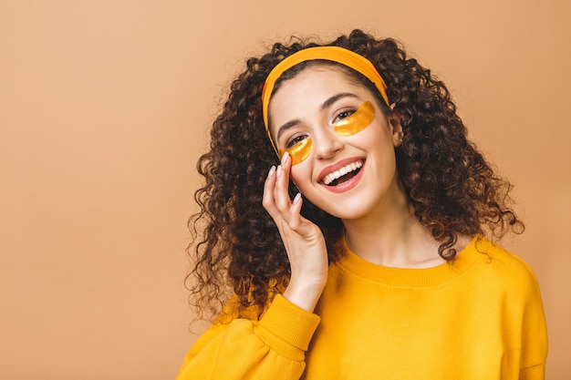 L'immagine di giovane bella donna riccia pura isolata sopra fondo beige si prende cura della sua pelle con le bende dell'occhio.