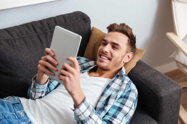 L'immagine di un allegro uomo vestito con una camicia in una gabbia stampata giace sul divano di casa e usa un tablet