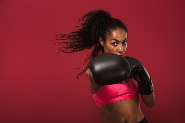 L'immagine di un fantastico giovane pugile africano sportivo in posa isolata sul muro rosso fa esercizi di boxe.