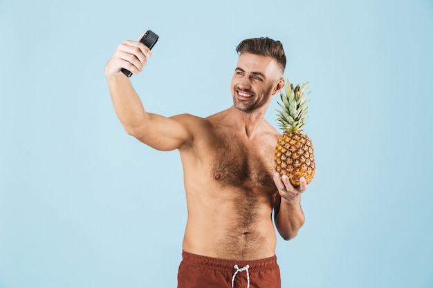 L'immagine di un uomo adulto felice eccitato bello che posa sopra la parete blu prende un selfie dal telefono che tiene l'ananas.