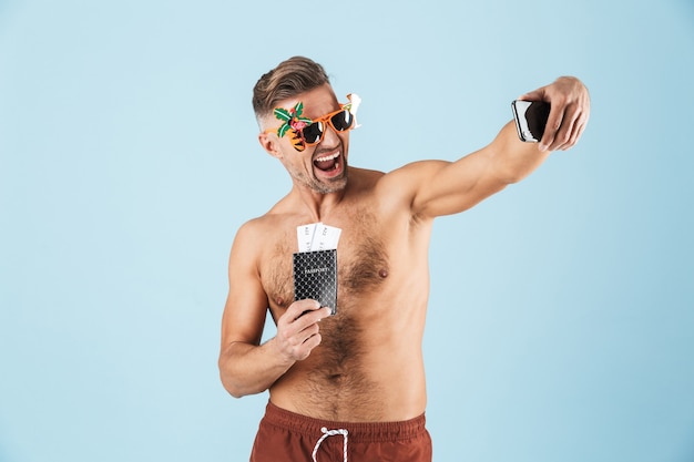L'immagine di un uomo adulto felice eccitato bello in costume da bagno che posa sopra la parete blu usando il telefono cellulare prende un passaporto della tenuta del selfie con i biglietti.
