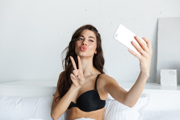 L'immagine di una bella donna castana che indossa lingerie utilizzando il telefono cellulare si trova a letto prendere un selfie che mostra il gesto di pace.