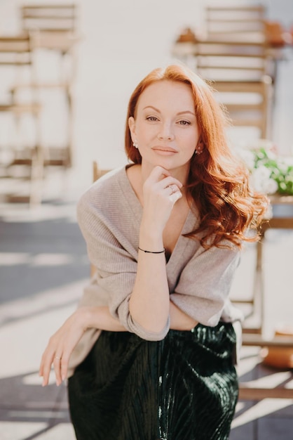 L'immagine di una bella donna con i capelli rossi ha il trucco tiene la mano sotto il mento guarda direttamente la fotocamera indossa abiti eleganti posa su sfondo di strada ha un'espressione sicura di sé Bellezza e riposo