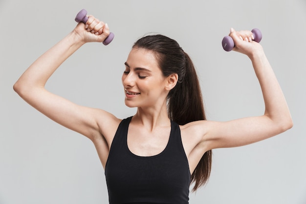 L'immagine di una bella giovane donna sportiva fitness fa esercizi con manubri isolati sul muro grigio.