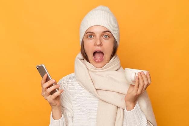 L'immagine di una donna caucasica stupita e sorpresa che indossa un berretto caldo e una sciarpa in posa isolata su sfondo giallo tiene la bocca spalancata tenendo in mano il cellulare