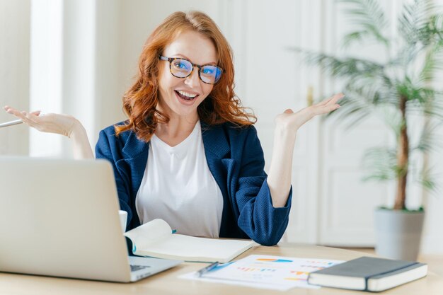 L'immagine di una donna d'affari dai capelli rossi allegra guarda il webinar o il video tutorial utilizza la connessione Internet gratuita alza i palmi delle mani con esitazione si siede sul desktop con i documenti Proprietaria di una piccola impresa