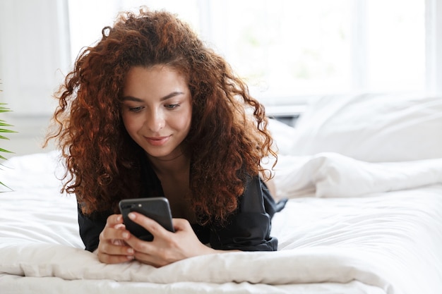 L'immagine di una donna soddisfatta in abito di seta della biancheria si trova in albergo domestico sul letto facendo uso del telefono cellulare.