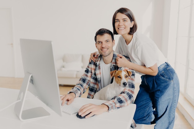 L'immagine di una donna sorridente bruna felice si appoggia alle spalle del marito che le tastiere sul computer navigano in Internet e cercano un nuovo appartamento da acquistare sul sito Web posano in una stanza spaziosa con il cane domestico